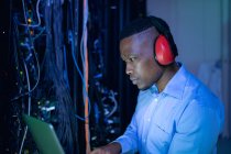 Afrikanisch-amerikanischer Computertechniker mit Kopfhörern und Laptop, der im Serverraum arbeitet. digitale Informationsspeicherung und Kommunikations-Netzwerktechnologie. — Stockfoto