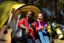 Happy diverse couple sitting in the tent and drinking coffee in countryside. healthy, active outdoor lifestyle and leisure time. — Stock Photo