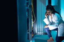 African american female computer technician making call and using laptop working in server room. digital information storage and communication network technology. — Stock Photo