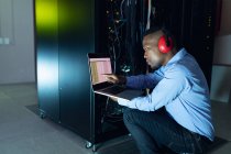 Africano americano técnico de computador masculino usando fones de ouvido usando laptop trabalhando na sala do servidor. armazenamento digital de informações e tecnologia de rede de comunicação. — Fotografia de Stock