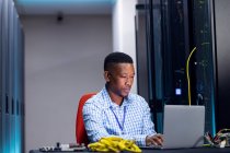 Sonriente afroamericano técnico de computación masculino utilizando portátil que trabaja en la sala de servidores de negocios. tecnología de redes digitales de almacenamiento y comunicación de información. - foto de stock