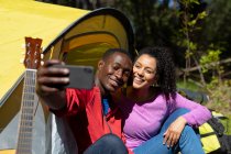 Felice coppia diversificata seduto nella tenda e prendendo selfie in campagna. stile di vita all'aperto sano e attivo e tempo libero. — Foto stock