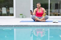Feliz afro-americano plus size mulher sentada no tapete e usando smartphone por piscina. fitness e estilo de vida saudável e ativo. — Fotografia de Stock