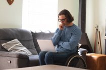 Задумчивый кавказский инвалид в очках, сидящий на инвалидном кресле с ноутбуком дома. Концепция инвалидности и инвалидности — стоковое фото