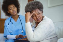 Doctora afroamericana hablando con una triste paciente mayor en casa. salud y estilo de vida durante la pandemia de covid 19. - foto de stock