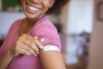 Mujer afroamericana sonriente que muestra vendaje en el brazo después de la vacunación covid. salud y estilo de vida durante la pandemia de covid 19. - foto de stock