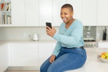 Heureuse femme afro-américaine plus taille ayant appel vidéo sur smartphone dans la cuisine. mode de vie, loisirs, passer du temps à la maison avec la technologie. — Photo de stock