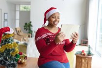 Feliz afro-americano plus size mulher em santa chapéu fazendo chamada de vídeo de Natal em tablet. Natal, festividade e tecnologia de comunicação. — Fotografia de Stock