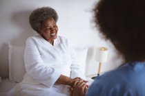 Afroamerikanische Ärztin hält lächelnde ältere Patientinnen zu Hause an den Händen. Gesundheitswesen und Lebensstil während der Covid 19 Pandemie. — Stockfoto