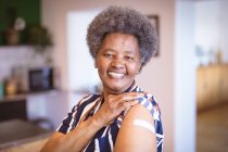 Porträt einer lächelnden afrikanisch-amerikanischen Seniorin, die einen Verband am Arm nach einer Impfung zeigt. Gesundheitswesen und Lebensstil während der Covid 19 Pandemie. — Stockfoto