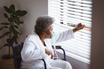 African american senior woman sitting on wheelchair with oxygen mask and looking through window. healthcare and lifestyle during covid 19 pandemic. — Stock Photo