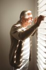 Senior caucasian man looking through window in his bedroom on sunny day. spending time at home alone. — Stock Photo