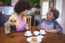 African american senior woman with adult daughter talking and drinking coffee in kitchen. family time at home together. — Stock Photo