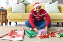 Happy african american plus size woman in santa hat wrapping presents at home. christmas, festivity and tradition concept. — Stock Photo