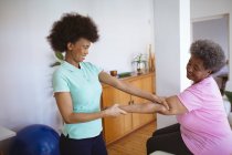 Sorrindo afro-americana fisioterapeuta do sexo feminino tratando braços de paciente sênior na clínica. cuidados de saúde seniores e tratamento de fisioterapia médica. — Fotografia de Stock
