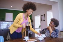 Smiling african american senior woman with adult daughter drinking coffee in kitchen. family time at home together. — Stock Photo