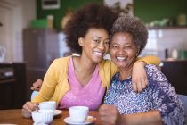 Retrato de una sonriente mujer mayor afroamericana con una hija adulta bebiendo café y abrazándose. tiempo en familia en casa juntos. - foto de stock