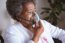 Donna anziana afroamericana seduta sulla sedia a rotelle con maschera di ossigeno a casa. assistenza sanitaria e stile di vita durante la pandemia della congrega 19. — Foto stock