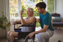 Zweirassiger männlicher Physiotherapeut behandelt Arme eines älteren männlichen Patienten im Rollstuhl in der Klinik. Senior Health und medizinische physiotherapeutische Behandlung. — Stockfoto