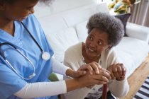 Lächelnde afrikanisch-amerikanische Ärztin an der Hand einer älteren Patientin zu Hause. Gesundheitswesen und Lebensstil während der Covid 19 Pandemie. — Stockfoto