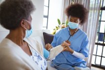 African american female doctor with face mask treating senior female patient at home. healthcare and lifestyle during covid 19 pandemic. — Stock Photo