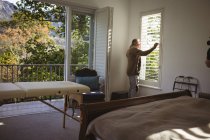 Hombre caucásico mayor mirando por la ventana de su dormitorio en un día soleado. pasar tiempo en casa solo. - foto de stock