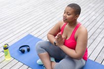 Afroamericano plus size donna in abiti sportivi seduto su tappetino e praticare yoga. fitness e stile di vita sano e attivo. — Foto stock