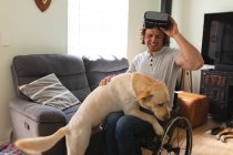 Homem caucasiano deficiente vestindo vr fone de ouvido sentado em cadeira de rodas tocando seu cão em casa. conceito de deficiência e deficiência — Fotografia de Stock
