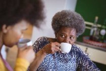 Mulher idosa afro-americana com filha adulta bebendo café na cozinha. tempo de família em casa juntos. — Fotografia de Stock