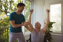 Smiling biracial fisioterapeuta masculino tratar braços de paciente do sexo masculino sênior em cadeira de rodas na clínica. cuidados de saúde seniores e tratamento de fisioterapia médica. — Fotografia de Stock