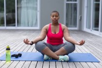 Mujer afroamericana de talla grande con ropa deportiva sentada en una esterilla y practicando yoga. fitness y estilo de vida saludable y activo. - foto de stock