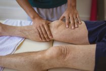 Бирациальный физиотерапевт-мужчина лечит ногу старшего пациента в клинике. медицинское и физиотерапевтическое лечение. — стоковое фото