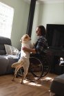 Белый инвалид, сидящий в инвалидном кресле и играющий дома со своей собакой. Концепция инвалидности и инвалидности — стоковое фото