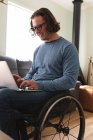 Uomo disabile caucasico che indossa gli occhiali seduto sulla sedia a rotelle utilizzando il computer portatile a casa. concetto di disabilità e handicap — Foto stock