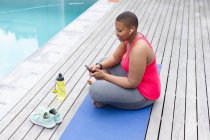 Happy african american plus size woman sitting on mat and using smartphone by swimming pool. fitness and healthy, active lifestyle. — Stock Photo
