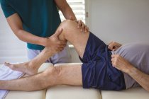 Zweirassiger männlicher Physiotherapeut behandelt Bein eines älteren männlichen Patienten in Klinik. Senior Health und medizinische physiotherapeutische Behandlung. — Stockfoto