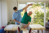 Biracial male physiotherapist with face mask treating arms of smiling senior male patient at clinic. senior healthcare and medical physiotherapy treatment. — Stock Photo