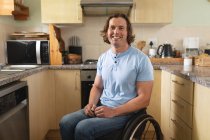 Retrato del hombre caucásico discapacitado sentado en silla de ruedas sonriendo en la cocina de su casa. concepto de discapacidad y discapacidad - foto de stock