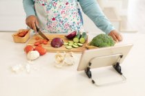 Midsection de plus taille femme coupant des légumes, en utilisant une tablette dans la cuisine. mode de vie, cuisine et passer du temps à la maison — Photo de stock