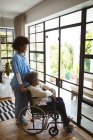 Smiling african american female doctor treating senior female patient on wheelchair at home. healthcare and lifestyle during covid 19 pandemic. — Stock Photo