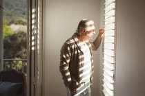 Senior caucasian man looking through window in his bedroom on sunny day. spending time at home alone. — Stock Photo
