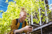 Hombre afroamericano albino feliz con rastas usando teléfono inteligente. nómada digital sobre la marcha, fuera y alrededor de la ciudad. - foto de stock