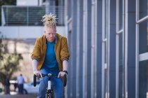 Pensativo albino hombre afroamericano con rastas montar en bicicleta. sobre la marcha, fuera y alrededor de la ciudad. - foto de stock