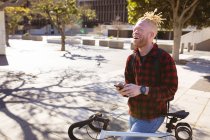 Happy albino african american man with dreadlocks on bike using smartphone. digital nomad on the go, out and about in the city. — Stock Photo