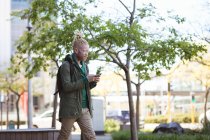 Homme américain albinos réfléchi avec dreadlocks marche à l'aide d'un smartphone. nomade numérique en déplacement, en déplacement dans la ville. — Photo de stock