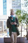 Homme afro-américain albinos avec masque facial et dreadlocks marchant et utilisant un smartphone. sur la route, dehors et dans la ville pendant la pandémie covid 19. — Photo de stock