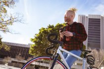Heureux albinos homme afro-américain avec dreadlocks sur vélo en utilisant un smartphone. nomade numérique en déplacement, en déplacement dans la ville. — Photo de stock