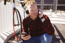 Hombre afroamericano albino feliz con rastas usando auriculares usando teléfono inteligente. nómada digital sobre la marcha, fuera y alrededor de la ciudad. - foto de stock