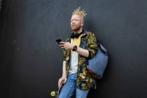 Homme américain albinos réfléchi avec dreadlocks tenant skateboard à l'aide d'un smartphone. nomade numérique en déplacement, en déplacement dans la ville. — Photo de stock