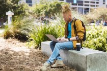 Homem americano albino atencioso afro-americano com dreadlocks sentado no parque usando laptop. nômade digital em movimento, para fora e sobre na cidade. — Fotografia de Stock